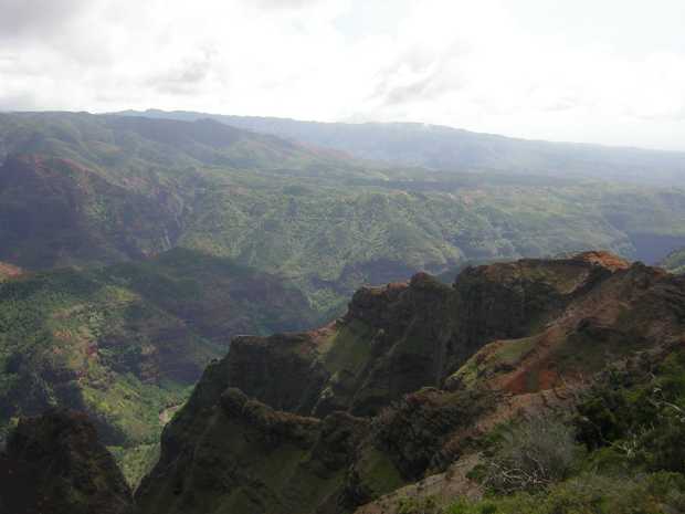 Beyond a rough dark red and green outcropping deep in shadow, folded green hills stretch out to the foggy horizon under a low gray sky. Beyond a rough dark red and green outcropping deep in shadow, folded green hills stretch out to the foggy horizon under a low gray sky.