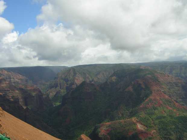 Dark red and gray craggy mountains, slathered here and there with green vegetation, stretch out below a flat horizon under a cloudy sky. Dark red and gray craggy mountains, slathered here and there with green vegetation, stretch out below a flat horizon under a cloudy sky.