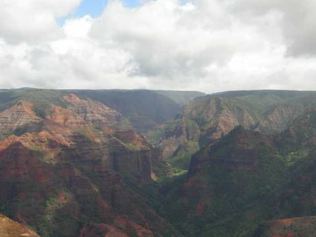 Horizontal layers in shades of red run across multiple triangular peaks zig-zagging into the distance; green vegetation climbs the lower parts of these canyon walls. Horizontal layers in shades of red run across multiple triangular peaks zig-zagging into the distance; green vegetation climbs the lower parts of these canyon walls.