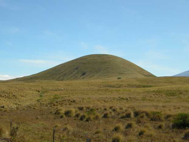 Against a nearly cloudless blue sky, an almost perfectly rounded, greenish tan hill rises suddenly from a nearly flat dry prairie of tufted grass.