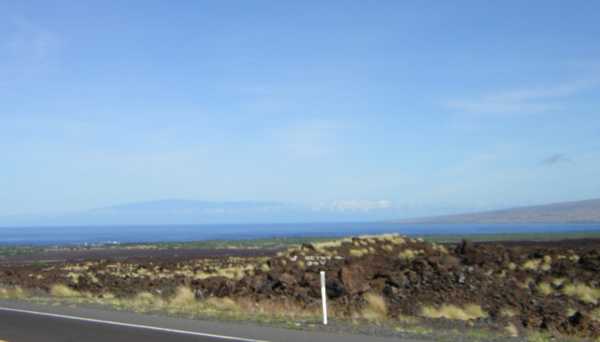 From a roadside, jagged reddish-brown rocky chunks interspersed with yellow-tan tufts of dry grass stretch away to the distant sea, beyond which a shadowy blue mountain rises.