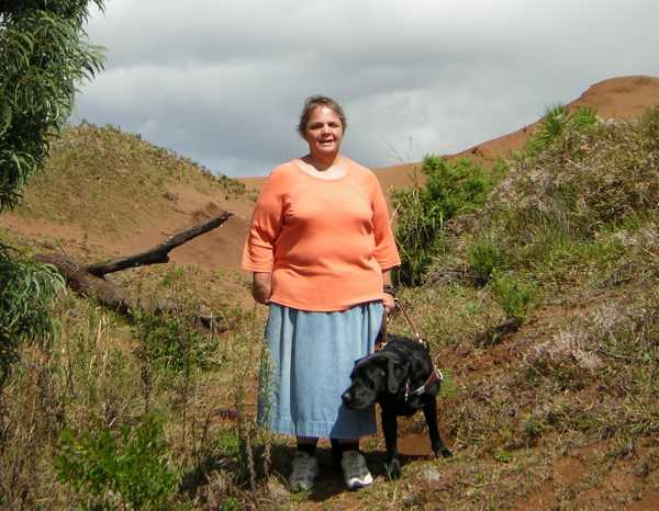 Maria, in orange shirt and blue denim skirt, and Bella, sniffing the foliage, stand amongst scrub plants before low mounds of red earth and a dead tree near Waimea Canyon Overlook Maria, in orange shirt and blue denim skirt, and Bella, sniffing the foliage, stand amongst scrub plants before low mounds of red earth and a dead tree near Waimea Canyon Overlook