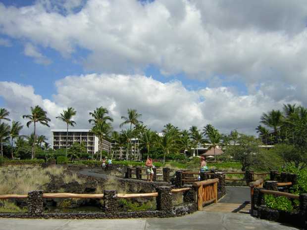 Waikoloa Beach Marriott: A white multi-story building among palm trees in the background; a zig-zag black paved path leads down through lava chunks and scrub grass, across a wooden bridge, to a concrete patio in the foreground.
