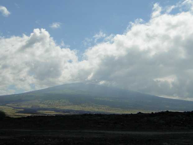 A distant, massively broad mountain rises from the prairie, its flank changing from tan desert to dark-green forest to a shadowed gray-blue bulk whose summit is hidden by cottony clouds.