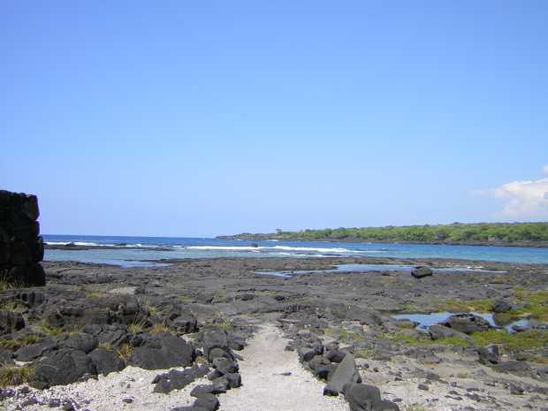 A white sandy path bordered by rough black cobbles leads away toward flat black lava and whitecapped surf beyond. The corner of a black stone wall is at left, and low forest extends out along the water at right.