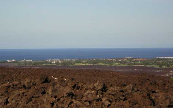 Looking down from a height, a reddish-brown jumble of rough rocks gives way to darker rock, then to a distant green swath of vegetation dotted with white and red buildings; beyond the sea stretches to the horizon.