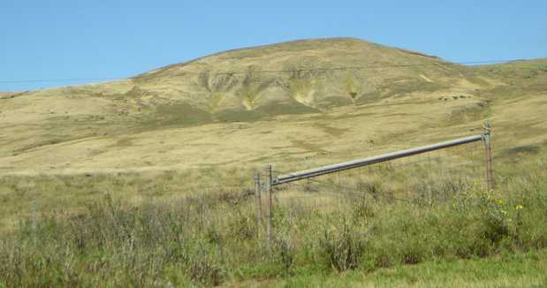 Behind a metal gate and barbed-wire fence, dry grass resembling light-tan velvet covers uneven prairie leading to a low hill whose flank is split by several deep vertical gullies.