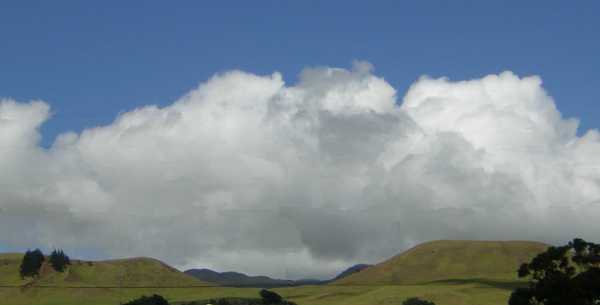 Velvety olive-green hills rise abruptly from a green plain against a backdrop of fluffy grey-white clouds topped with deep blue sky.