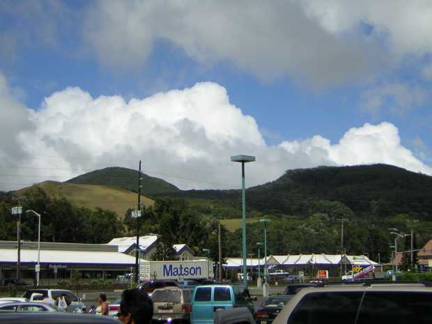 Beyond a parking lot filled with cars, vans, and people, a tractor-trailer bearing the name 'Matson', a McDonalds, and other buildings, stand against a tumble of pale green grassy, and dark green forested, hills.