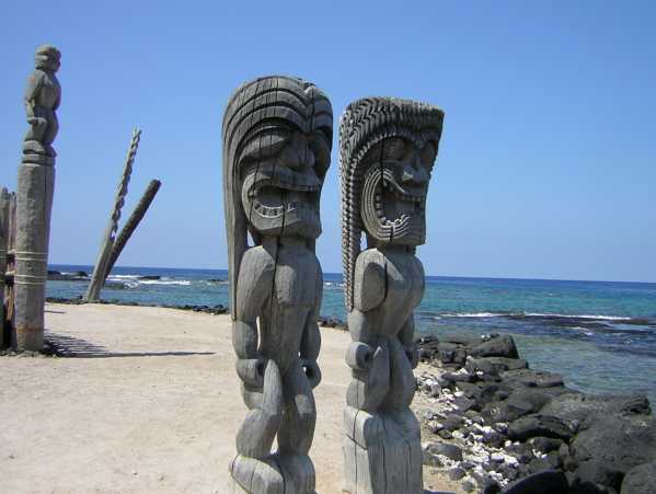 Two 9-foot grey carved wooden statues of humanoids with long hair and snarling mouths full of teeth stand on white sand near a seawall of black rocks; other carved statues and poles stand nearby.